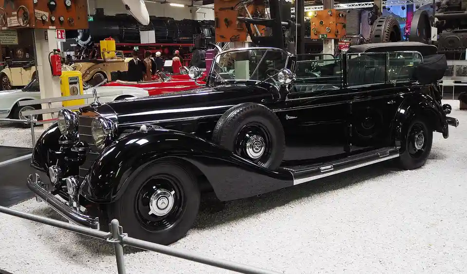 A black vintage Mercedes-Benz convertible is displayed in a museum, featuring a spare tire mounted on its side. The car is…