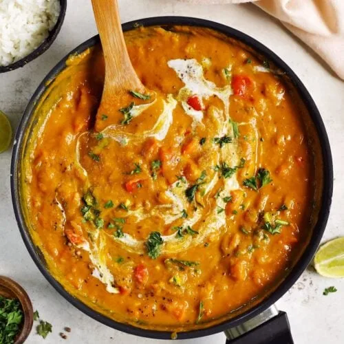 An overhead photo of a creamy orange curry in a black skillet, garnished with chopped cilantro and a swirl of cream, with a…