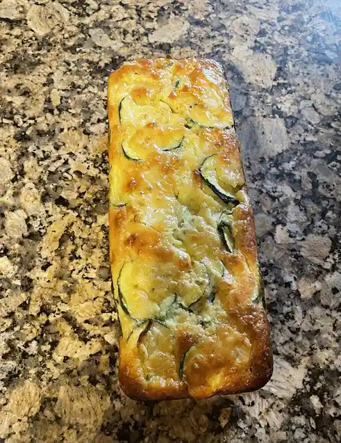 A golden-brown loaf of zucchini bread with visible green zucchini slices baked into the top sits on a speckled granite…