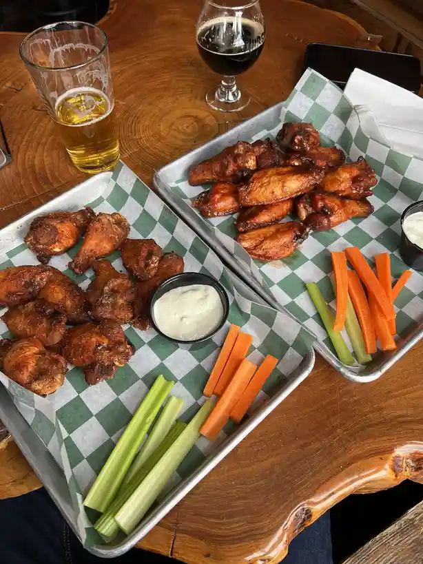 Two metal trays lined with green-and-white checkered paper hold chicken wings, carrot sticks, celery sticks, and small bowls…