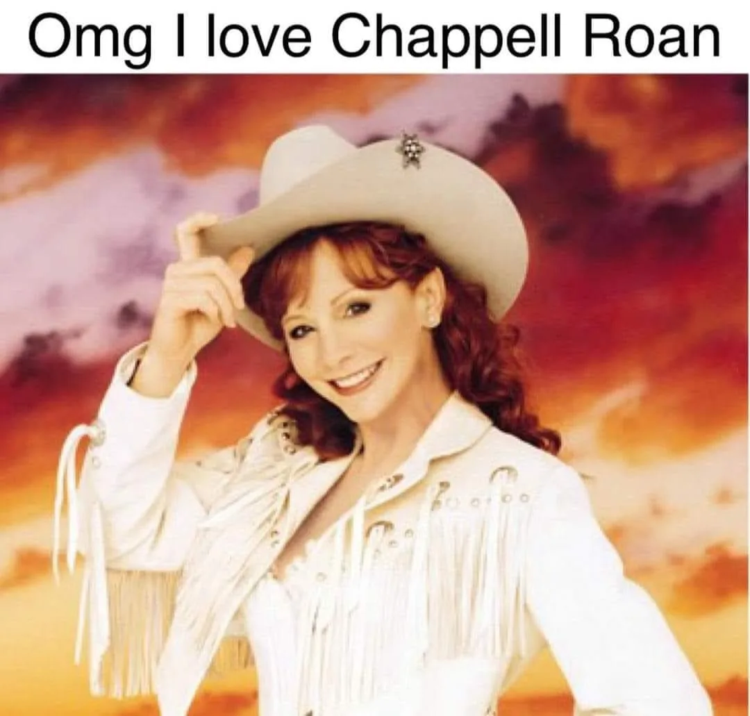 A woman with red hair wearing a white cowboy hat and a white fringed western-style jacket smiles while tipping her hat…