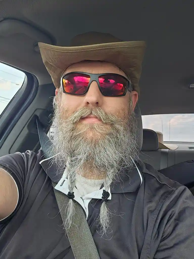 A man with a long, gray beard styled in two braids sits in a car wearing a brown cowboy hat and red-tinted sunglasses. He is…
