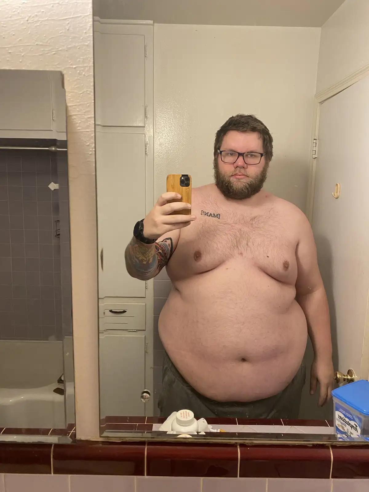 A shirtless, overweight man with a beard and glasses takes a mirror selfie in a bathroom, holding a phone with a wooden…