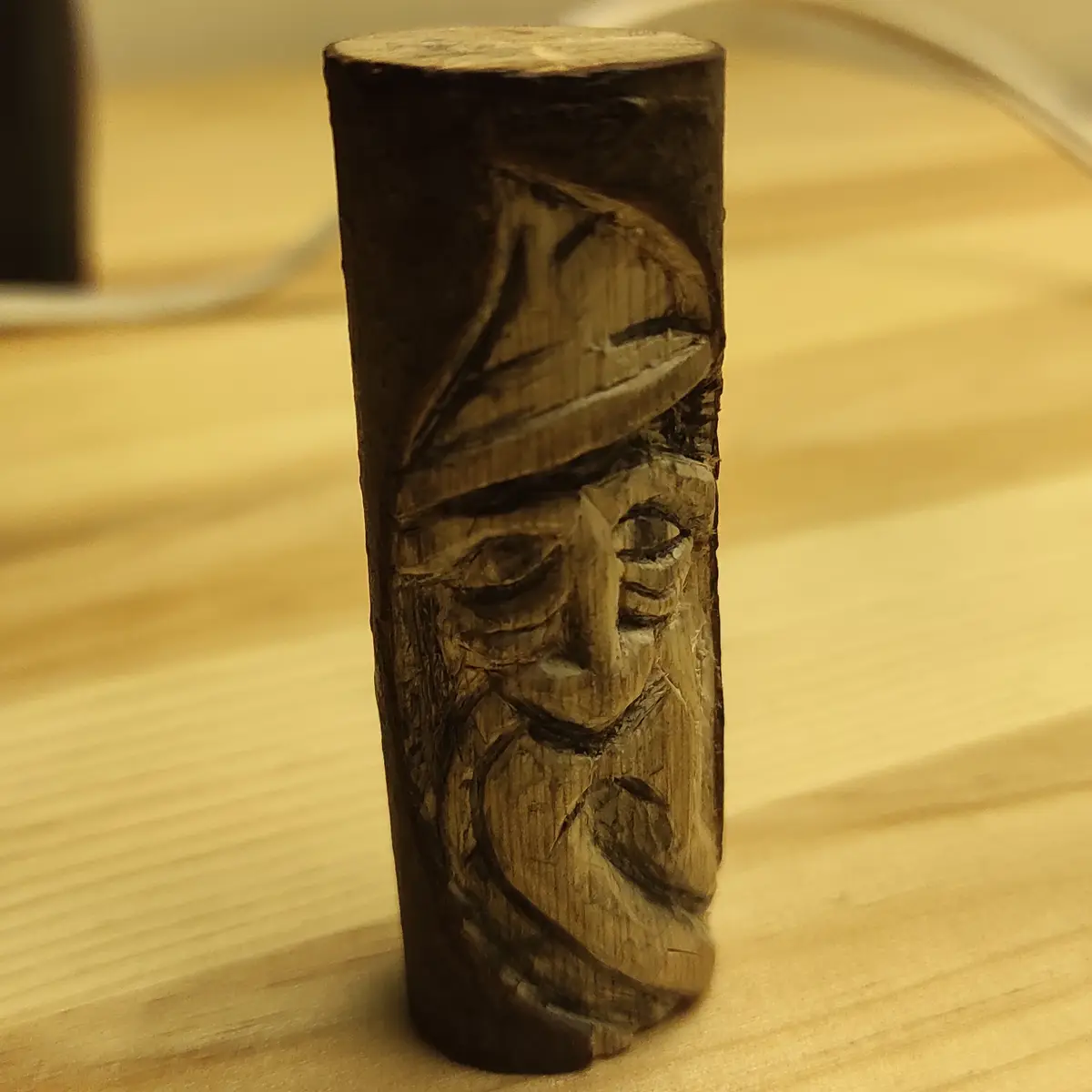 A close-up photo shows a small, hand-carved wooden cylinder standing upright on a light-colored wooden surface. The cylinder…