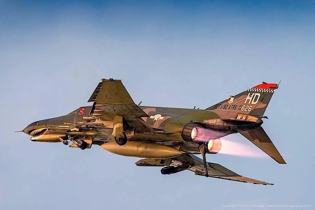 A military jet fighter, painted with a shark mouth design on the nose, is shown in flight against a clear blue sky, with its…