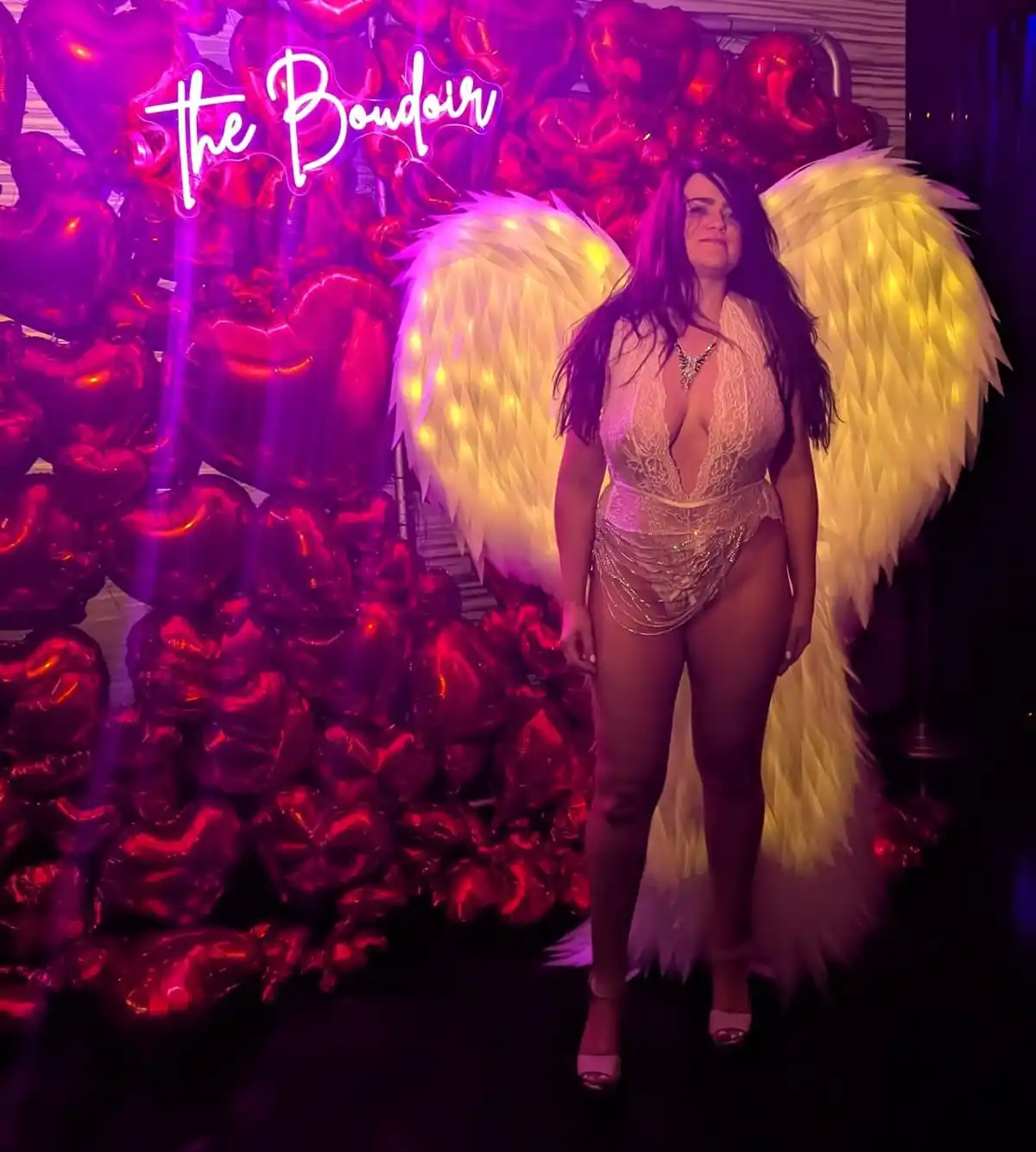 A woman with long dark hair stands in front of a wall of shiny red heart-shaped balloons under purple lighting, wearing a…