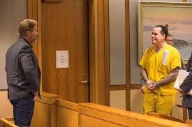 A man in a yellow prison jumpsuit stands in a courtroom, looking at another man in a suit who is leaning against a wooden…