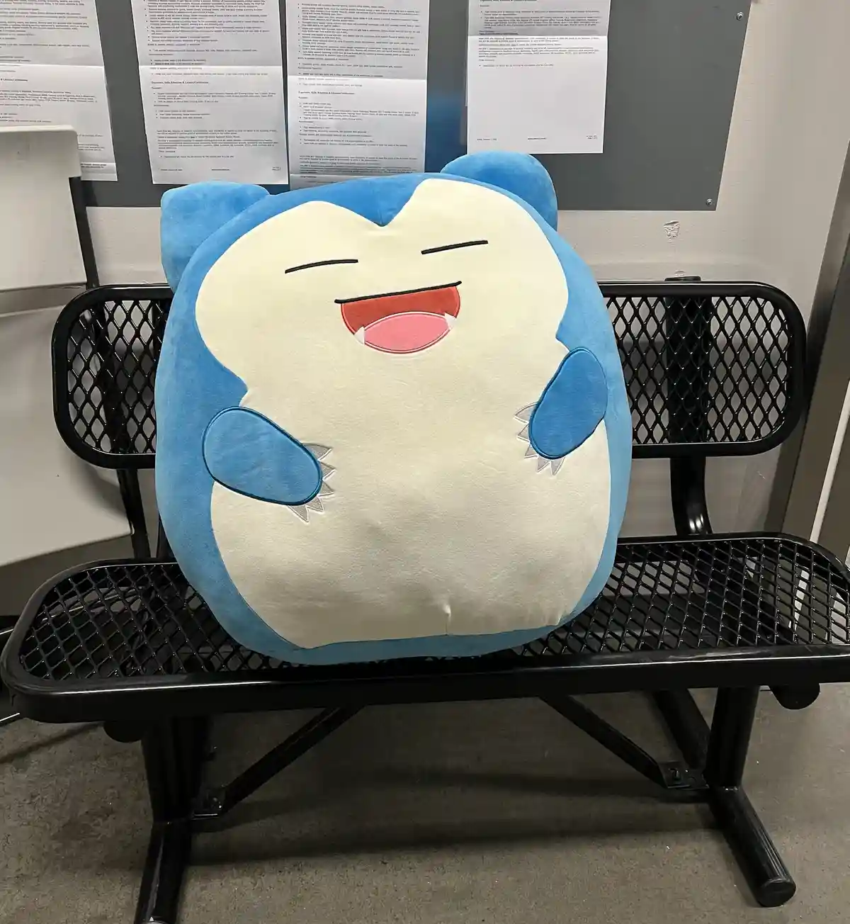 A large, round, blue and white plush toy resembling the Pokémon Wobbuffet sits on a black metal bench in what appears to be…