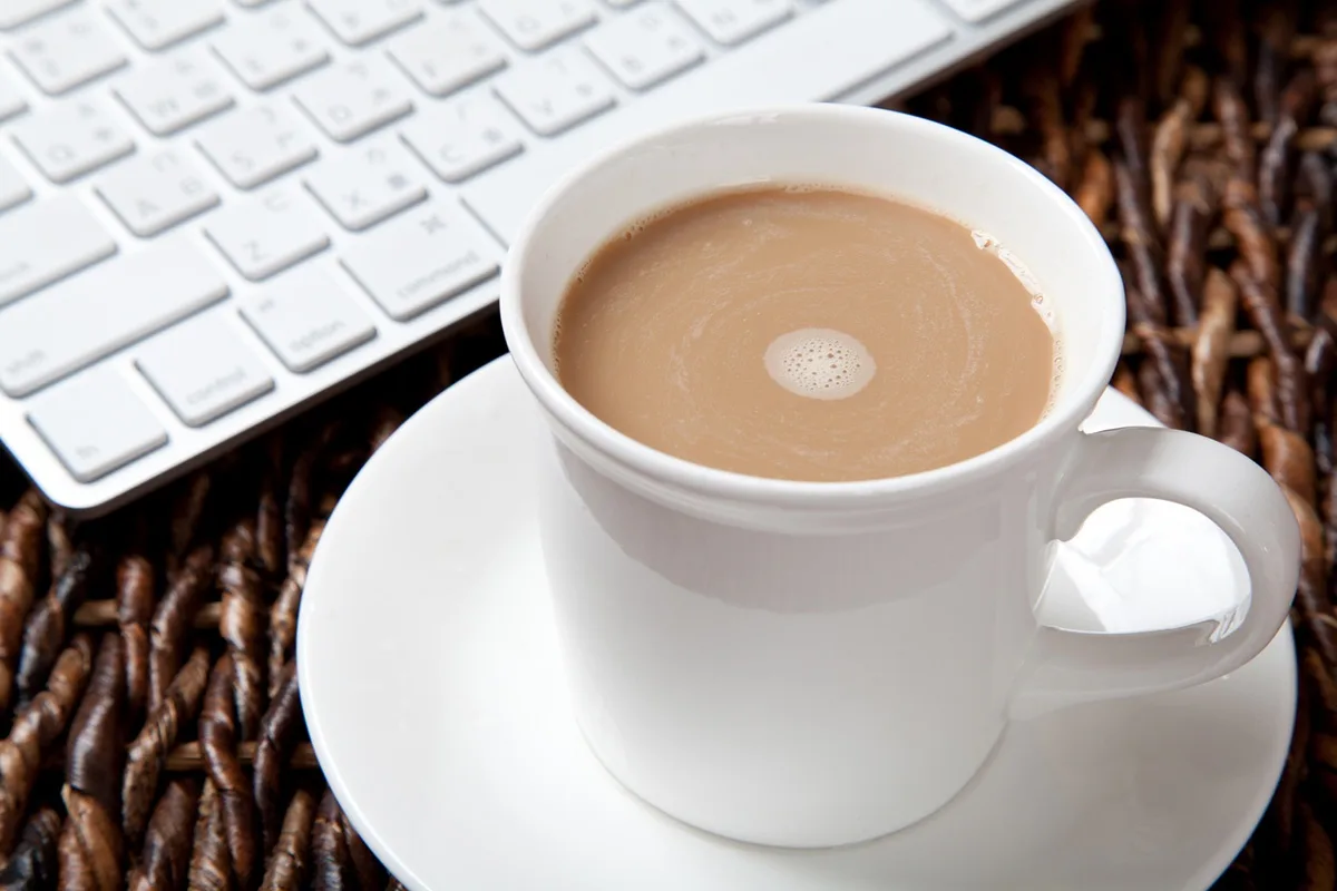 A white ceramic mug filled with light brown liquid, likely tea or coffee, sits on a matching saucer atop a dark woven mat. A…
