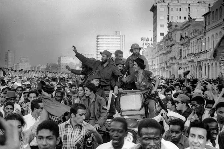 A black-and-white historical photograph shows Fidel Castro and Che Guevara waving from the back of a jeep as they ride…