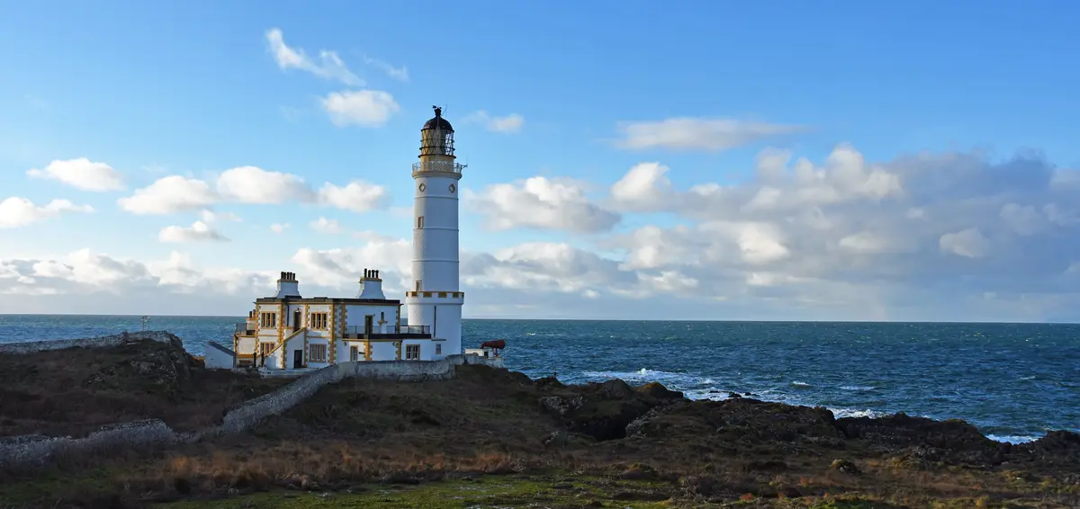 A scenic photo of a white lighthouse with a yellow-trimmed keeper's house on a rocky cliff overlooking the ocean under a…