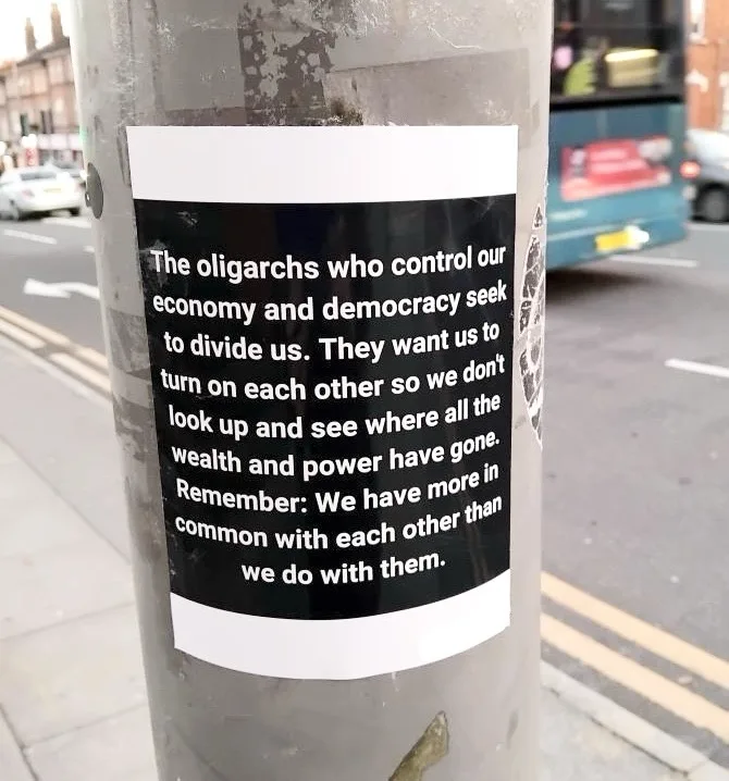 A black-and-white sticker is affixed to a grey metal utility pole on a city sidewalk, with a street, cars, and a bus visible…
