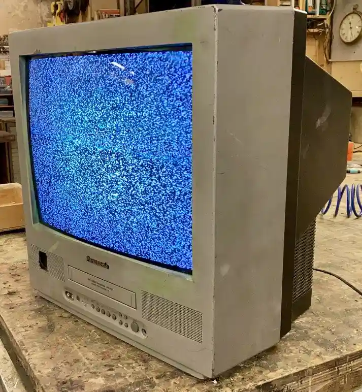 An old, bulky Panasonic CRT television sits on a cluttered workbench, its screen filled with blue static noise. The TV has…