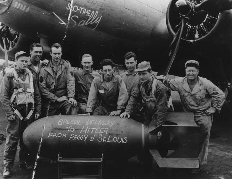 A black-and-white historical photo shows eight smiling U.S. airmen posing with a large bomb. The bomb has handwritten text…