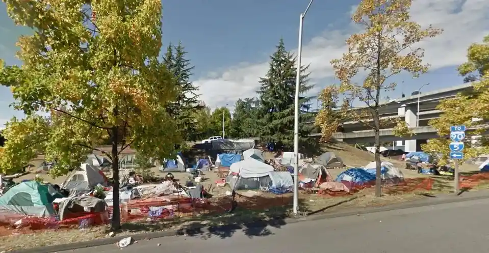 An outdoor area with numerous tents and makeshift shelters set up along a roadside, bordered by orange safety fencing and…