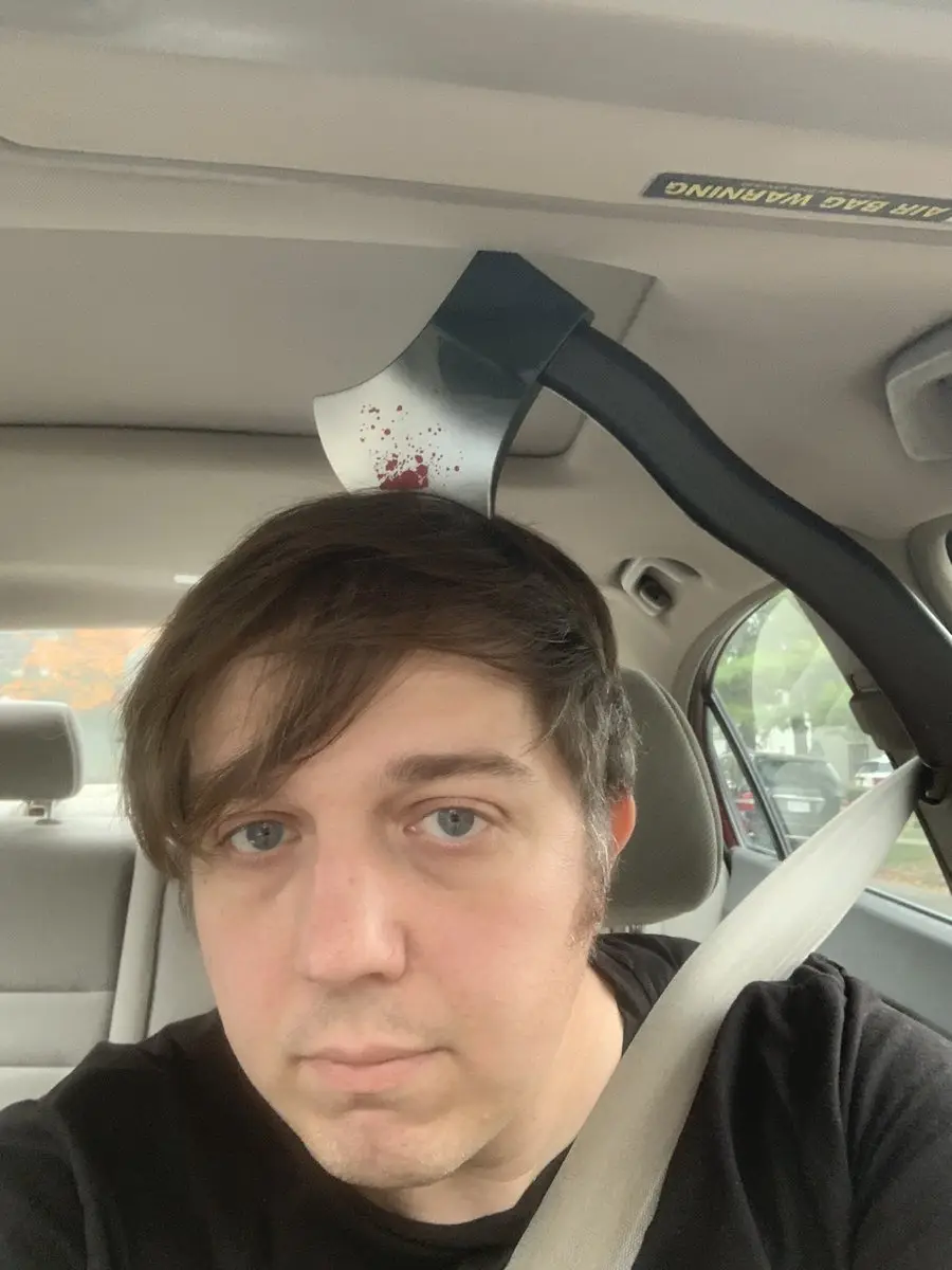 A man with brown hair and blue eyes sits in a car seat, looking directly at the camera with a neutral expression. An axe…