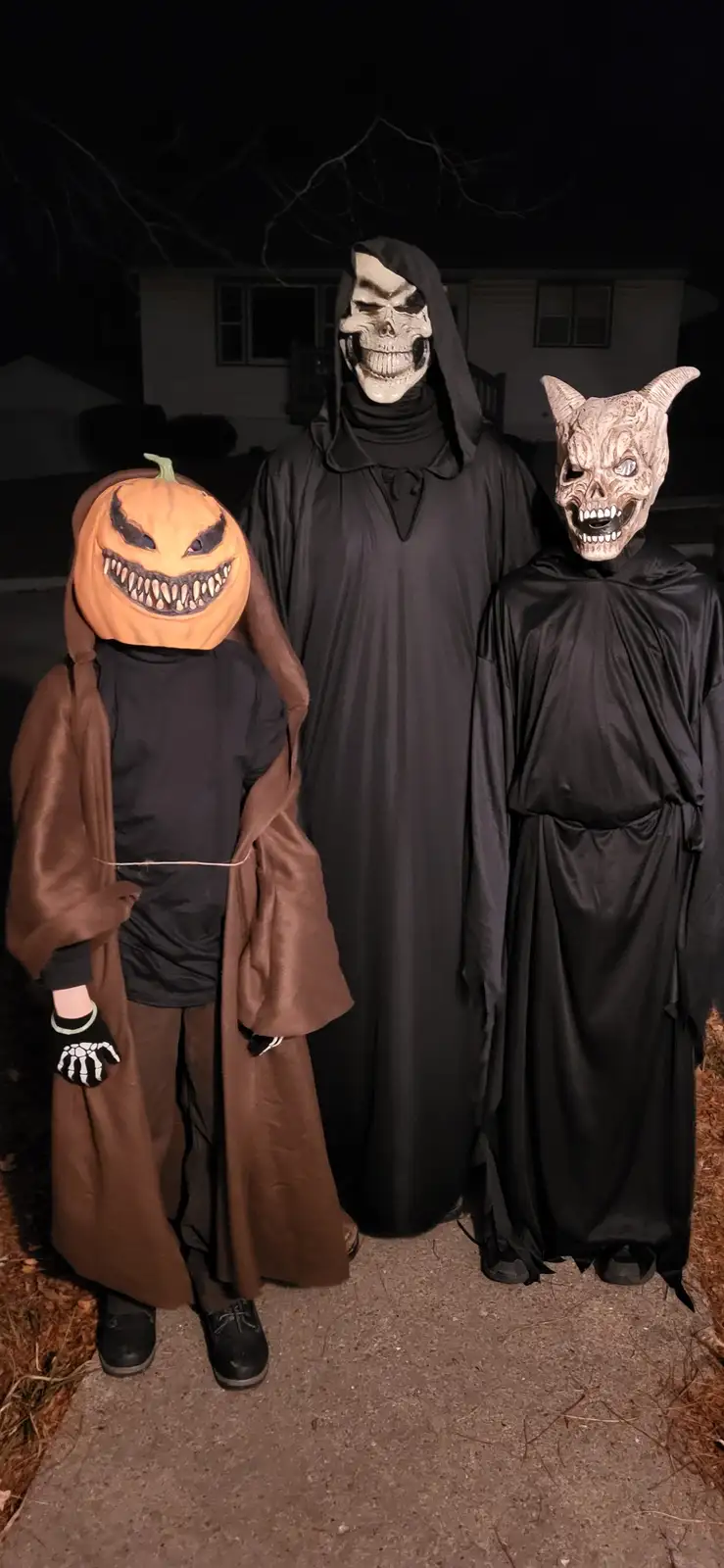 Three people in Halloween costumes stand outside at night: one wears a pumpkin head with a brown robe, another wears a skull…