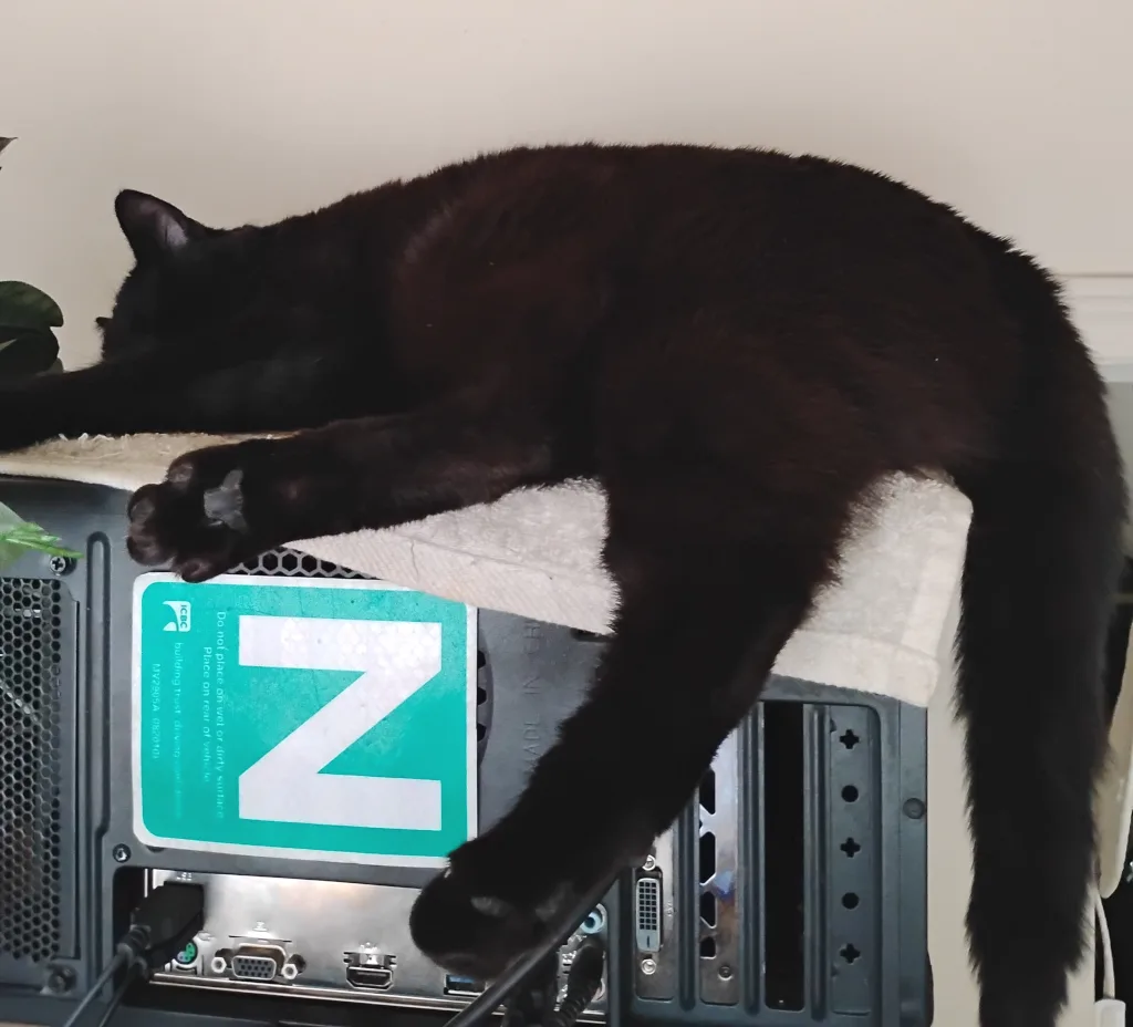 A black cat is stretched out and sleeping soundly on top of a computer tower, its body draped over a white towel. A large…