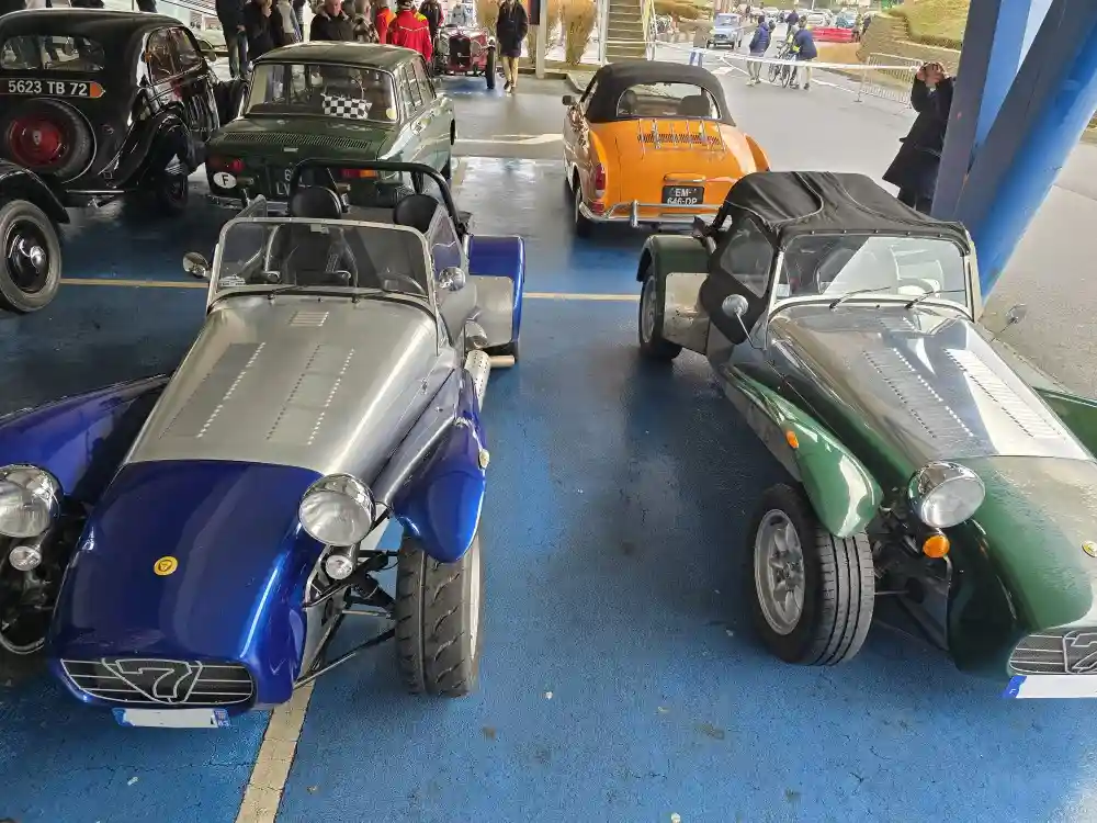 Two vintage-style open-top sports cars, one blue and one green, are parked side-by-side on a blue asphalt surface at what…