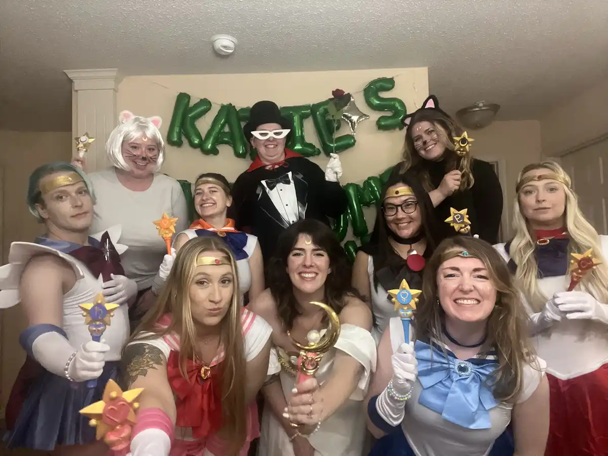 A group of friends dressed as characters from Sailor Moon, holding wands and posing for a photo in front of green balloon…