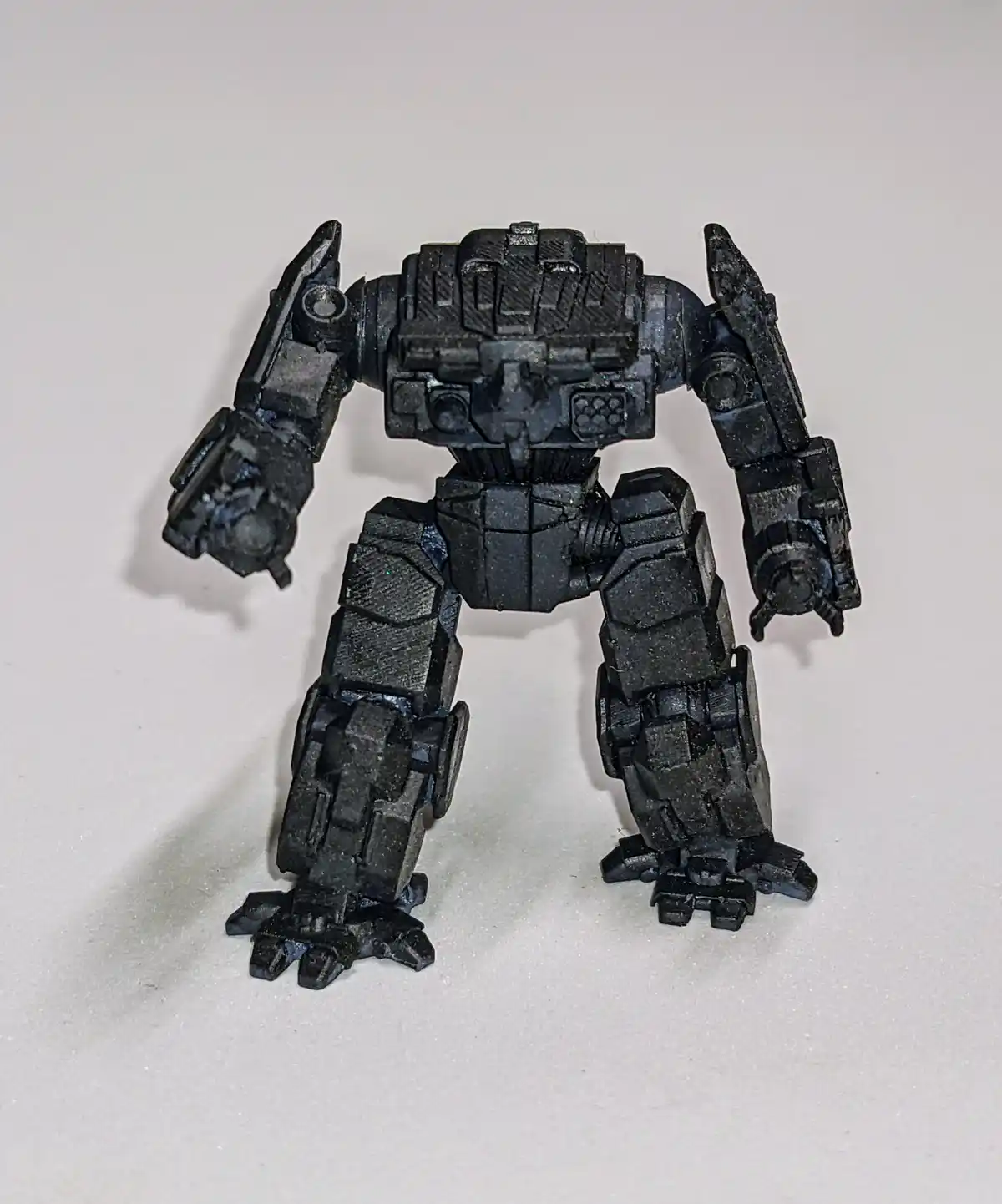 A black, heavily textured, 3D-printed humanoid robot figurine stands on a plain white surface, its arms slightly raised. The…