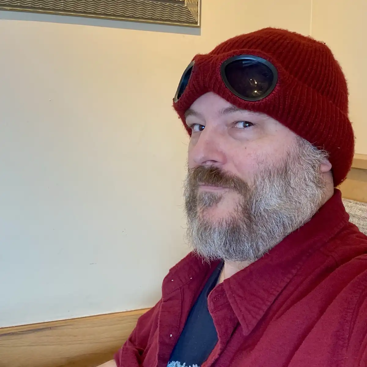 A man with a graying beard and mustache wears a red knit beanie with aviator-style goggles perched on top, looking directly…