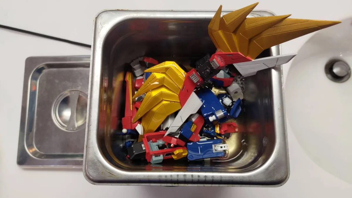 A stainless steel sink basin filled with disassembled, colorful plastic robot parts, mostly red, blue, and gold, submerged…