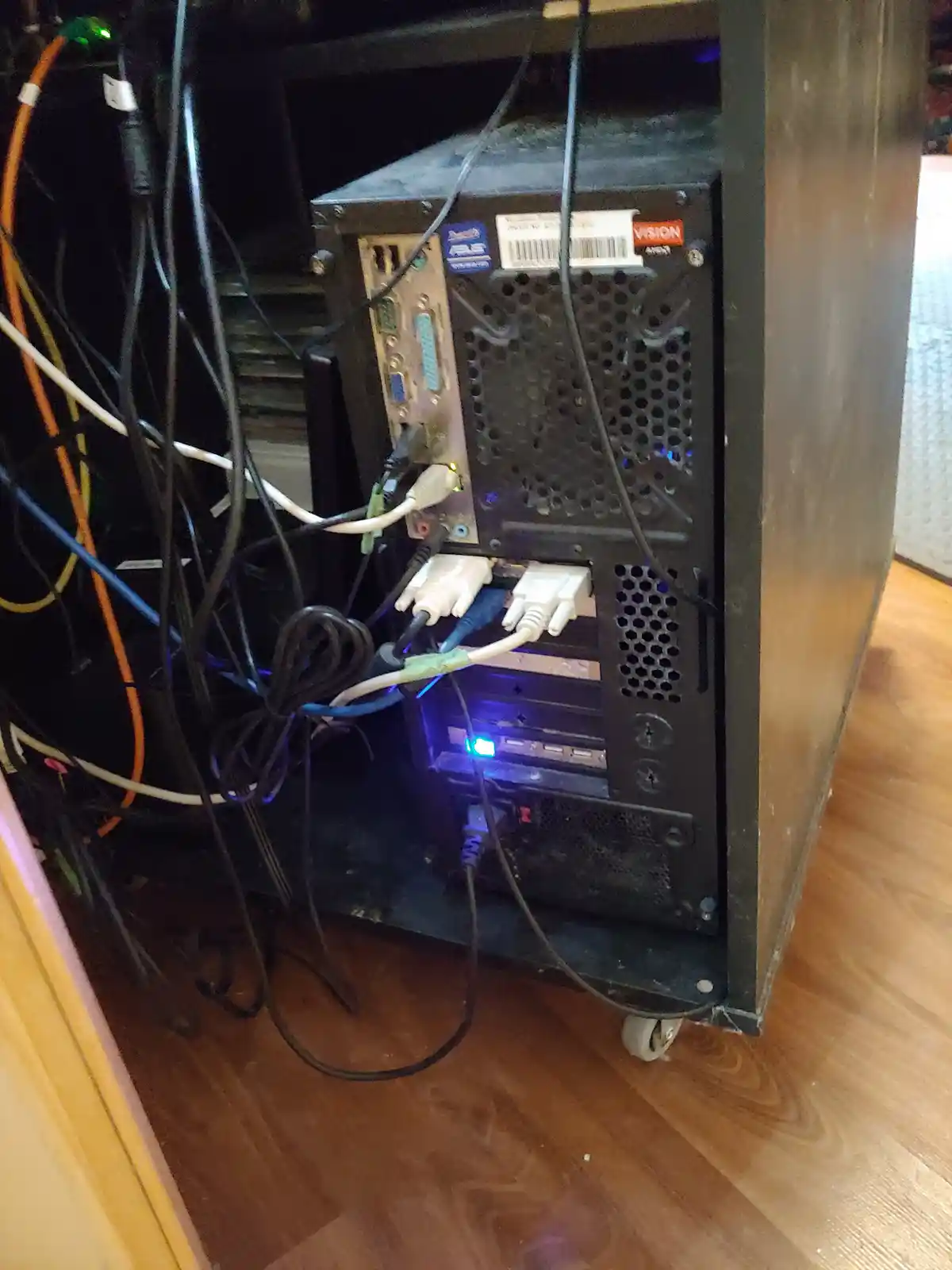 The image shows the back of a black computer tower with numerous tangled cables plugged into various ports, including VGA…