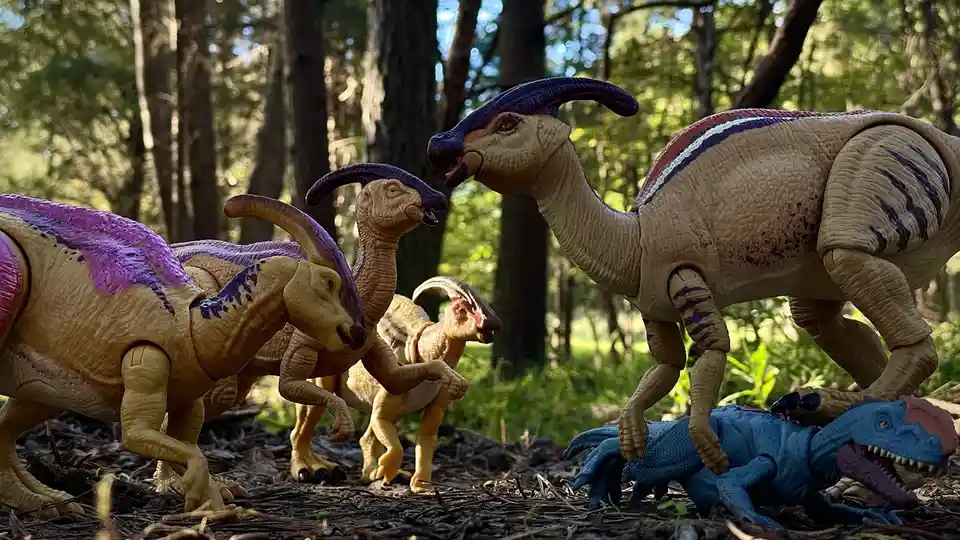 Several toy parasaurolophus dinosaurs with purple crests are staged in a forest, one standing over a blue toy dinosaur lying…