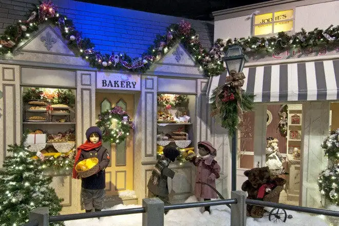 A festive, snow-covered diorama depicts a bakery and candy shop decorated with garlands and lights. Mannequins dressed in…