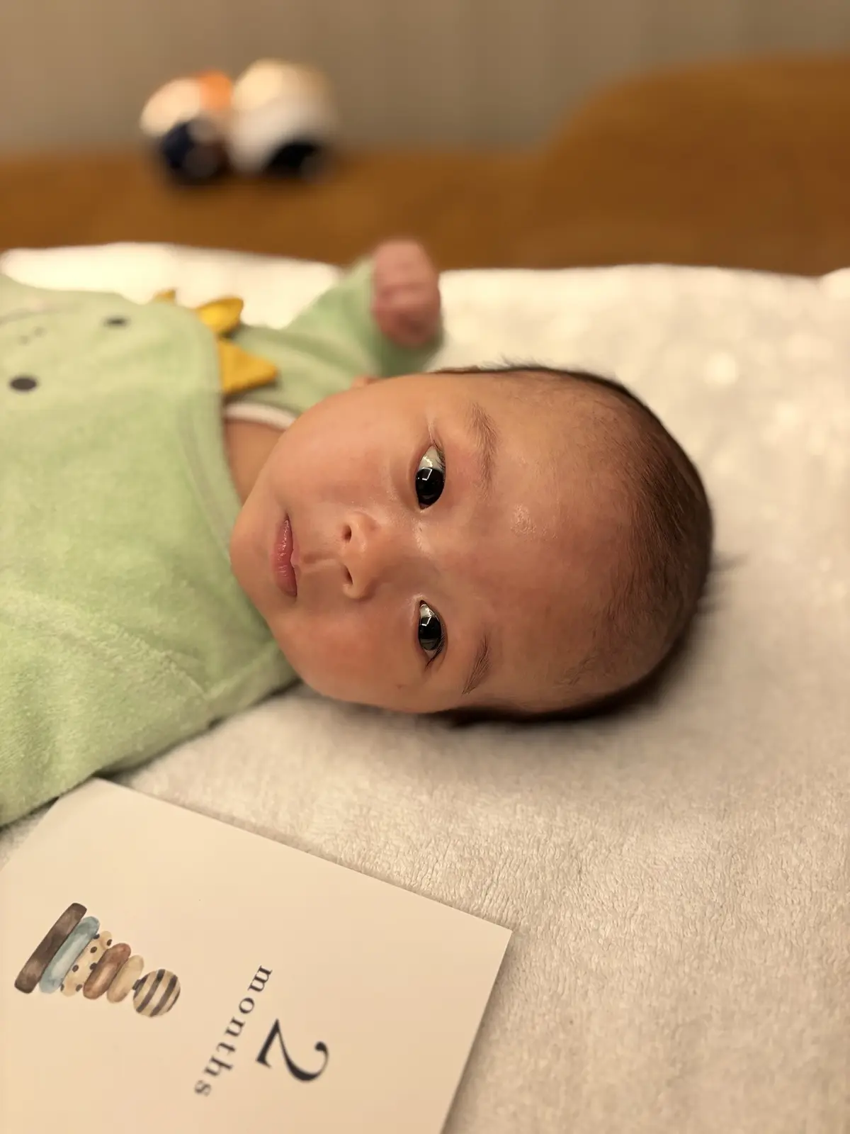 A close-up photo of a baby lying on its back on a white textured blanket, wearing a green onesie with a yellow dinosaur…