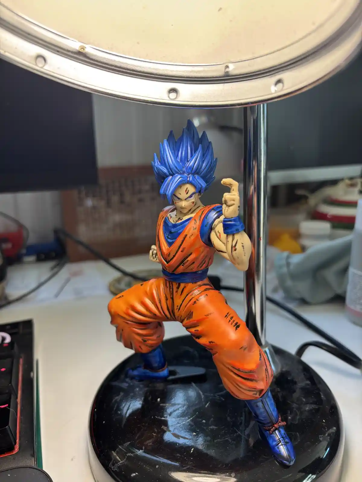 A blue-haired, orange-outfit Dragon Ball Z figurine of Goku in a battle stance stands on a black base under a desk lamp…