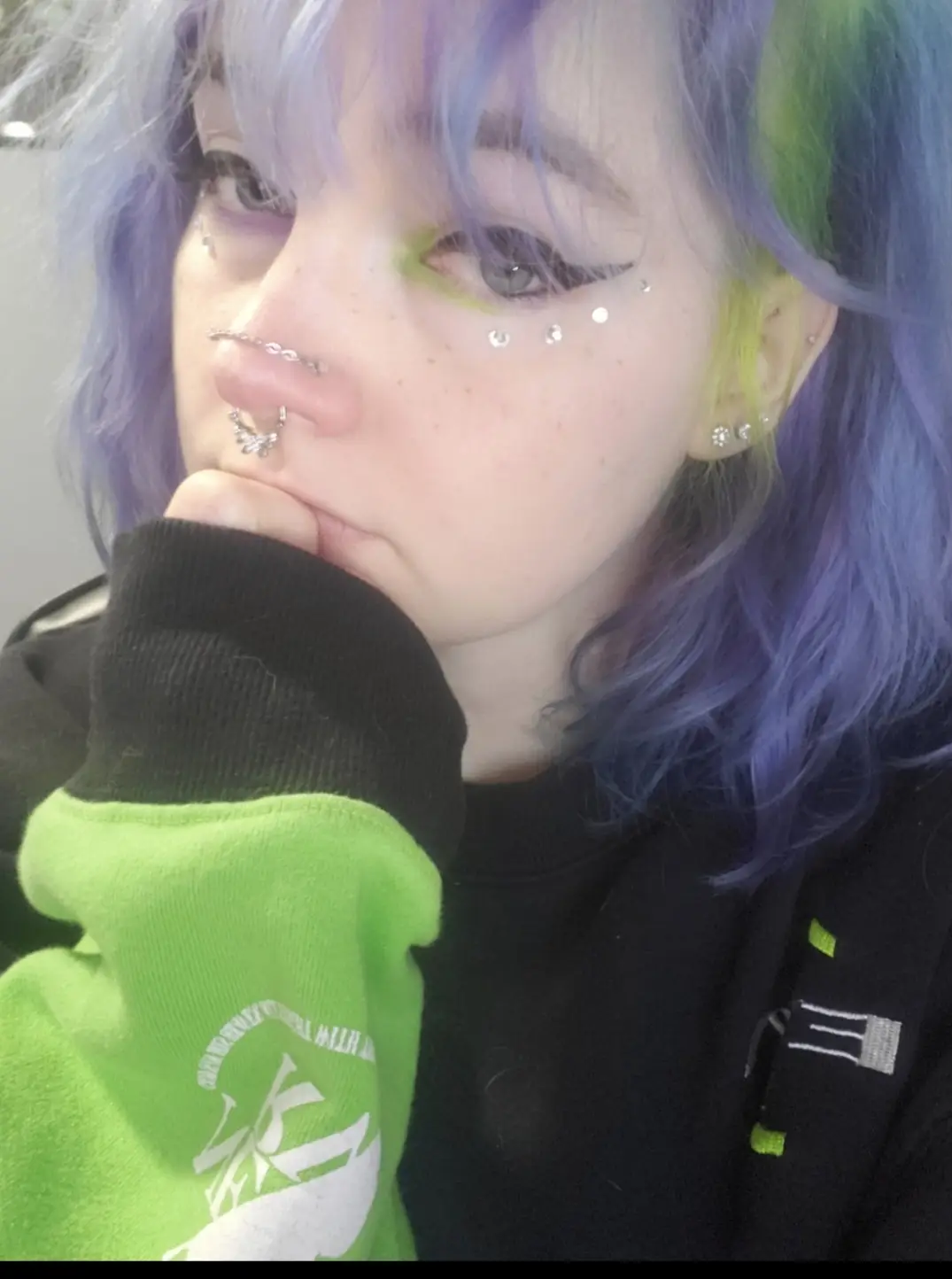 A person with vibrant purple and green hair, multiple facial piercings including a septum ring and cheek gems, and bold…