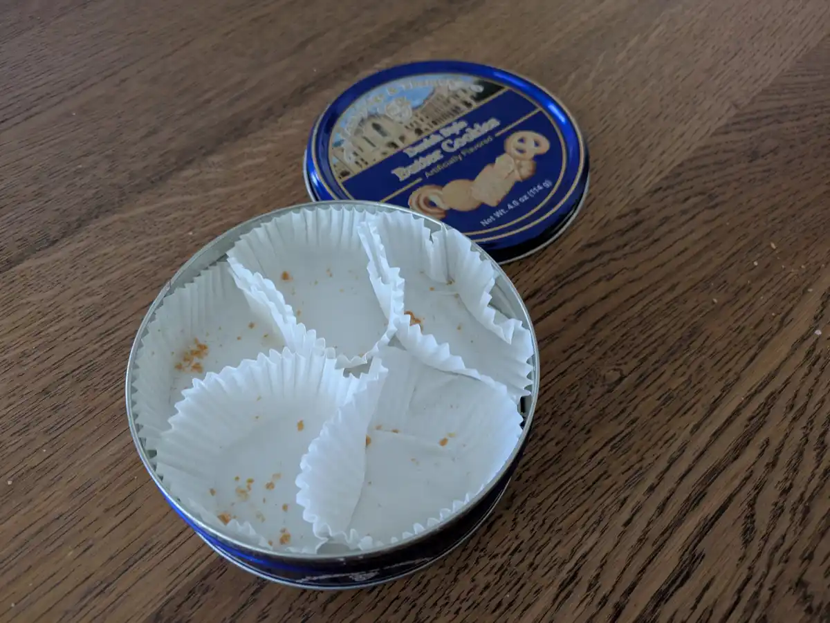 An open, empty blue tin of 'Cambridge & Thames Danish Style Butter Cookies' sits on a wooden table, its white paper cupcake…