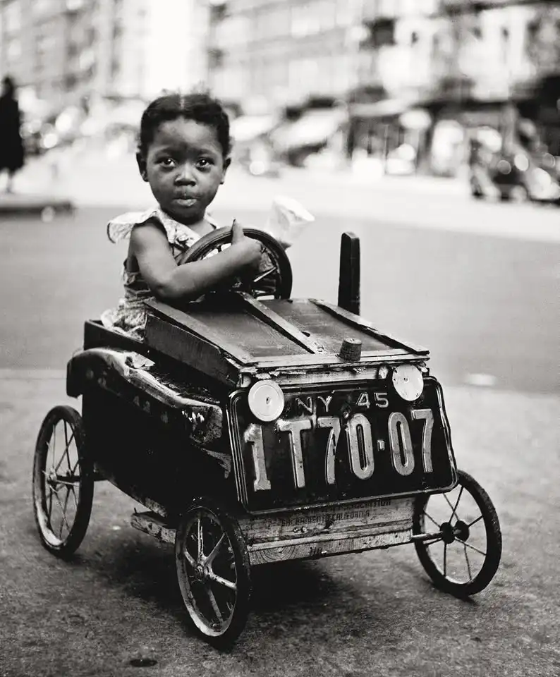 A black-and-white photo shows a young child sitting in a small, toy car with a New York license plate reading '1T70-07'. The…