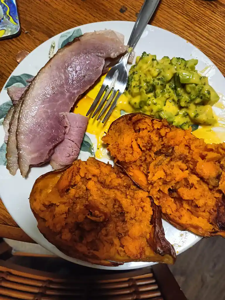 A plate of food featuring sliced ham, broccoli with cheese sauce, and two halves of a roasted sweet potato, with a fork…