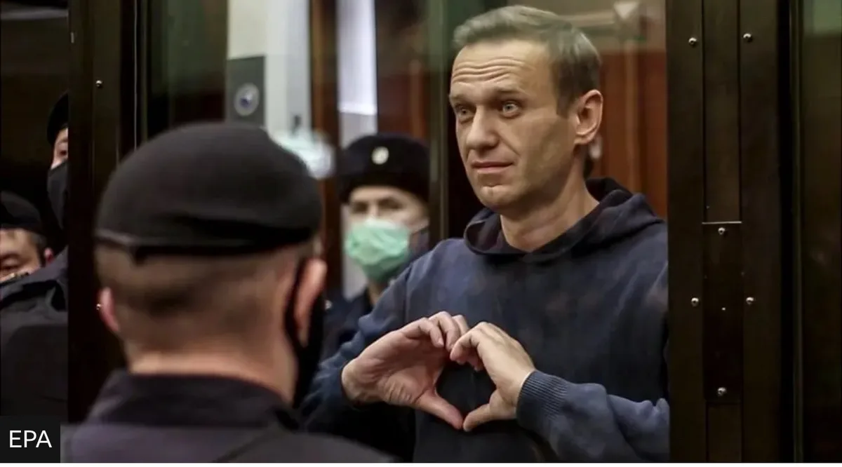 a man in a dark hoodie stands behind glass, forming a heart shape with his hands while looking at someone in front of him…