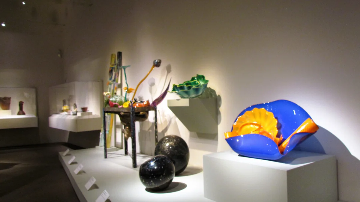 A museum gallery displays colorful, abstract glass sculptures on white pedestals under spotlights, including a large blue…
