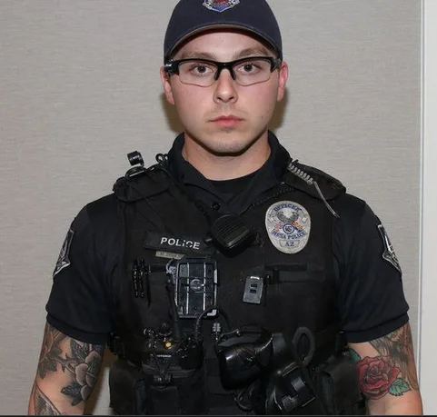 A young police officer in full uniform, including a cap, glasses, and tactical vest with 'POLICE' text, stands facing the…