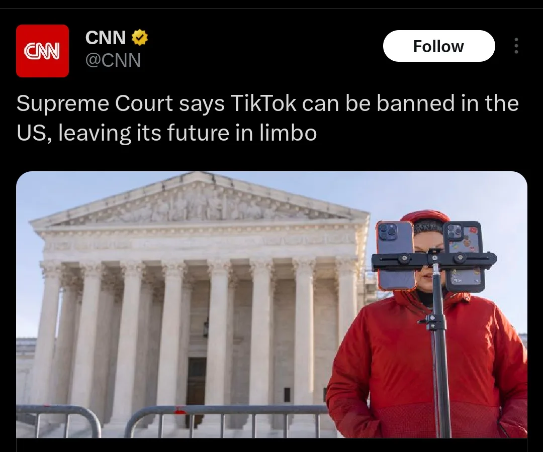 A person in a red jacket stands in front of the Supreme Court building, holding a selfie stick with two smartphones mounted…