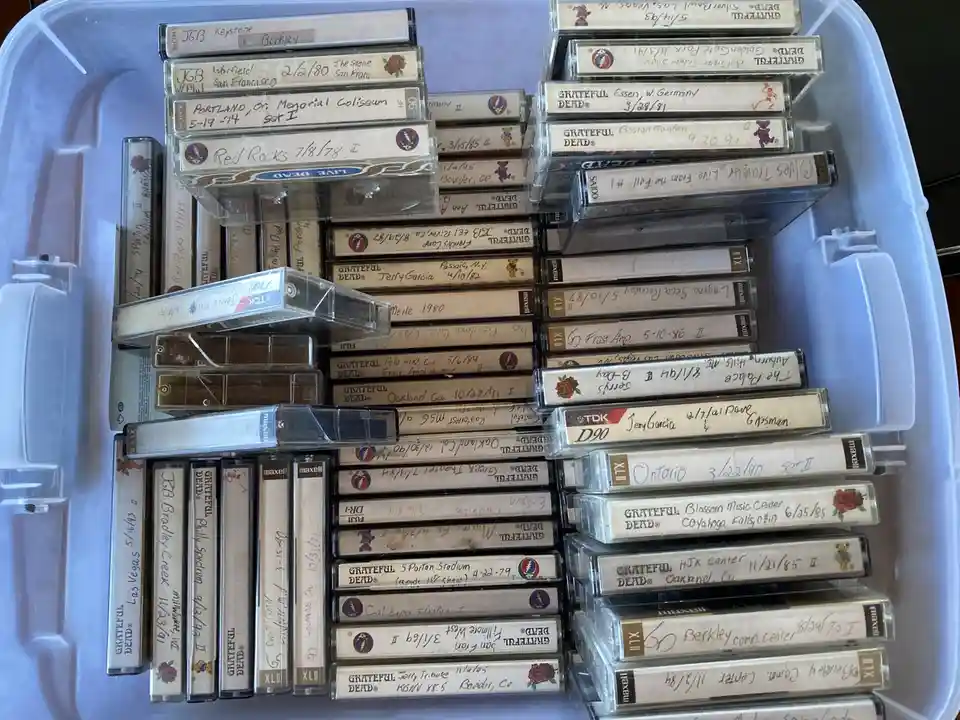 A light blue plastic storage bin is filled with dozens of cassette tapes, mostly labeled 'Grateful Dead' with handwritten…