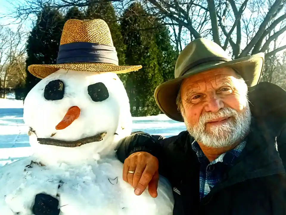 An older man with a white beard and a wide-brimmed hat poses for a selfie with a snowman he built, which also wears a straw…