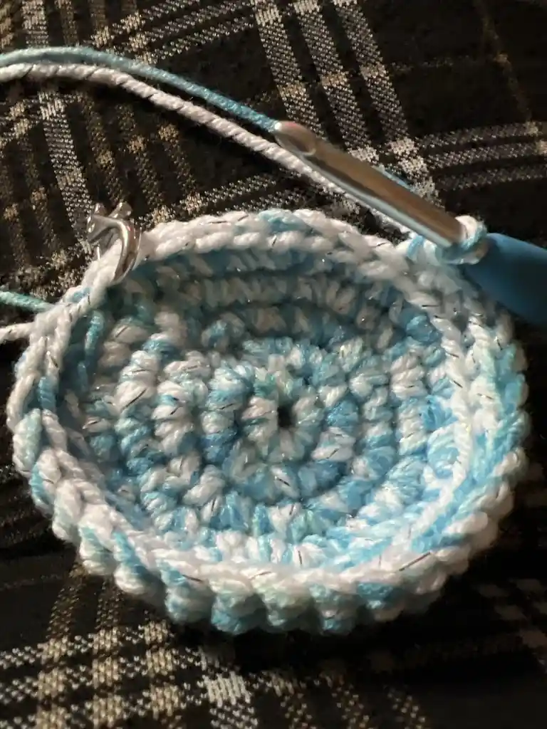 A close-up photo shows a partially completed circular crochet project made with light blue and white yarn, resting on a dark…