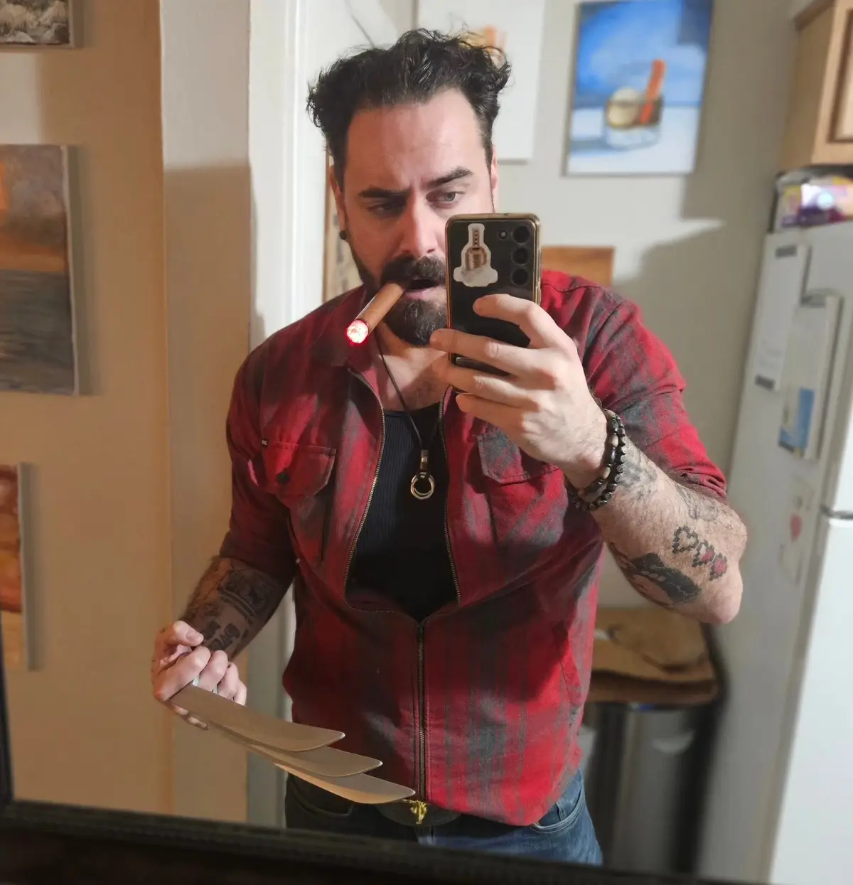 A man with a beard and tattoos takes a mirror selfie while holding a lit cigar in his mouth and three large kitchen knives…