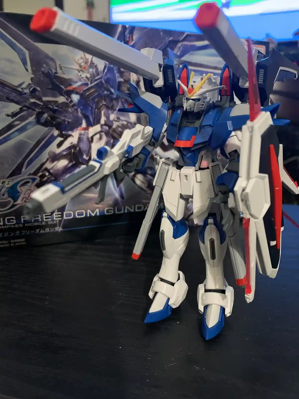 A blue, white, and red plastic Gundam model stands on a dark surface, holding two large weapons, with its box visible behind…