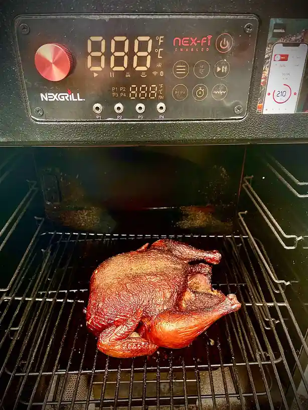 A roasted chicken sits on a rack inside a NexGrill smoker, which displays '888°F' on its digital panel, while a smartphone…