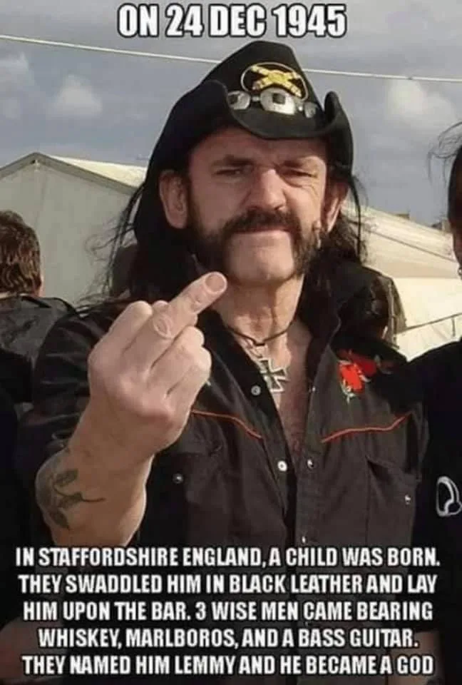 A man with long dark hair, a mustache, and a black leather jacket is pointing his middle finger at the camera. He wears a…