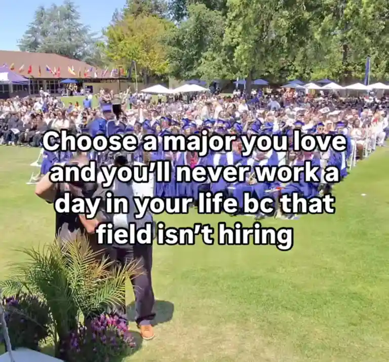 An outdoor graduation ceremony with many students in blue caps and gowns seated in rows on a grassy field. A person in a…