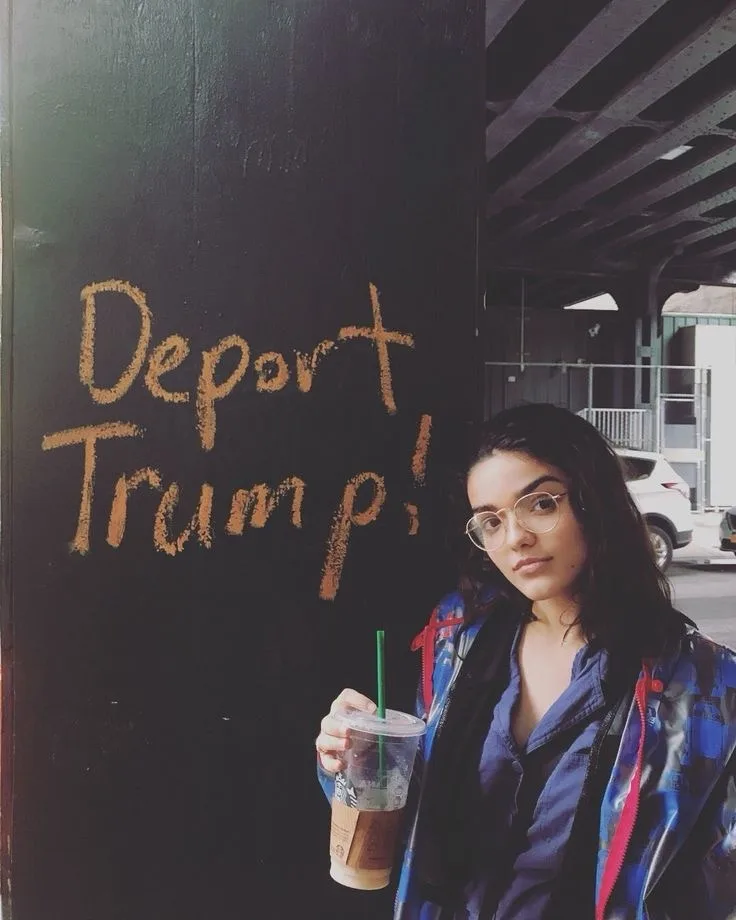 A young woman with dark hair and round glasses, wearing a blue jacket with red zipper details, holds a Starbucks iced coffee…