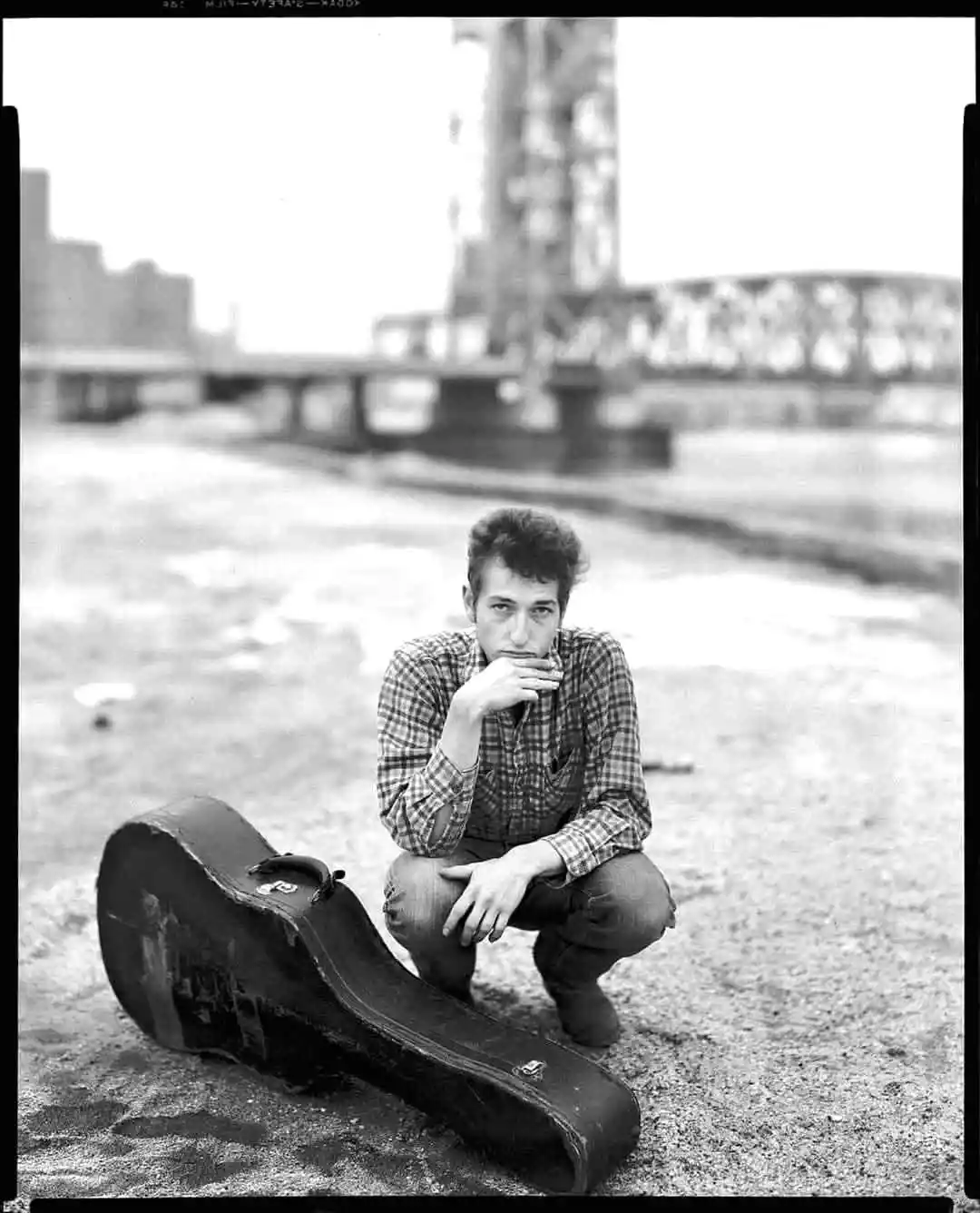 a black and white photo of a young bob dylan crouching beside an empty guitar case on a riverbank with a bridge in the…